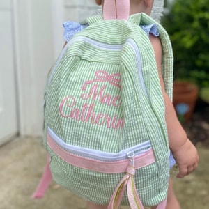May include: A green and white gingham backpack with a pink zipper and pink bow embroidery. The backpack is personalized with the name "Mac Catherine" in a pink script font.