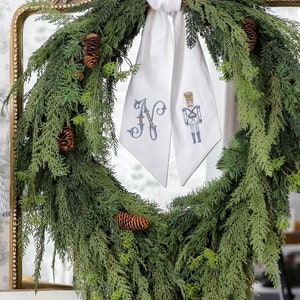 May include: A green pine wreath with pine cones hangs from a gold ornate mirror. A white ribbon with an embroidered letter "N" and a nutcracker design is tied around the wreath.