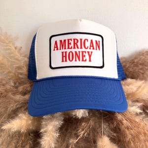 May include: A trucker hat with a white front panel and a blue brim and mesh back. The front panel features the words "AMERICAN HONEY" in red letters inside a black-bordered rectangle. The hat is displayed against a neutral background.