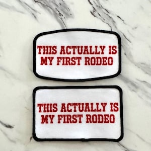 May include: Two rectangular patches with the text "THIS ACTUALLY IS MY FIRST RODEO" in red, embroidered on a white background. Each patch has a black border. The patches are displayed on a marble surface.
