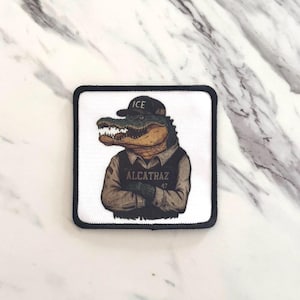 May include: Square patch featuring an alligator wearing a black cap with "ICE" on it, and a vest with "ALCATRAZ 47". The alligator has its arms crossed. The patch has a black border and is on a white background.