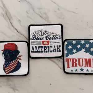 May include: Three embroidered patches with American patriotic themes. The first patch features a black labrador wearing a red, white, and blue bandana and a red baseball cap with the words "Make America Great Again". The second patch features a mountain range with the words "Blue Collar American" and "Dirty Hands Clean Money". The third patch features a blue and white American flag with white stars and the word "TRUMP" in red letters.