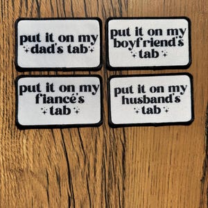 May include: Four rectangular white patches with black borders. Each patch has the phrase "put it on my" followed by "dad's tab", "boyfriend's tab", "fiancé's tab", and "husband's tab" in black text. Small star graphics are also present.