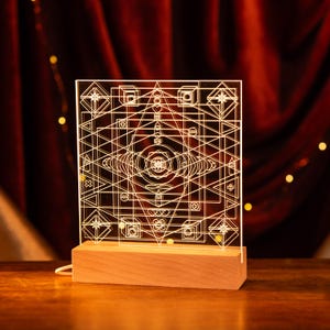 Op de afbeelding: Een geometrisch ontwerp wordt verlicht in een heldere acryl lamp met een houten basis. Het ontwerp heeft een verscheidenheid aan vormen en lijnen, waardoor een complex en ingewikkeld patroon ontstaat.