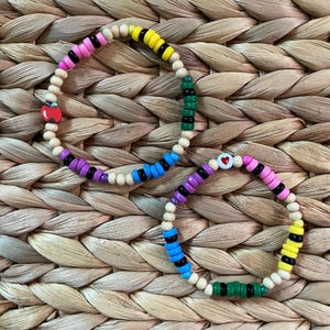 May include: Two colorful beaded bracelets with wooden beads, black beads, and heart-shaped charms. One bracelet has a red heart charm and the other has a pink heart charm.