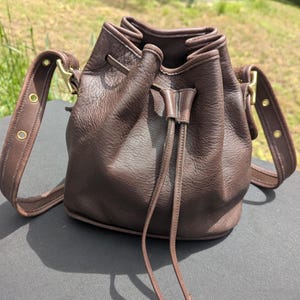 May include: A dark brown leather bucket bag with a drawstring closure, featuring a pebbled texture, a long adjustable shoulder strap with brass-toned metal hardware, and two long leather pull cords hanging from the front of the bag.