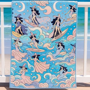 A beach towel with a whimsical design features illustrated witches surfing and floating on waves. The witches wear flowing dresses and wide-brimmed hats in pastel pinks, purples, and blues. The background is filled with swirling ocean waves, crescent moons, and stars, all in soft, dreamy colors. The towel hangs over a white railing with a view of the ocean and sandy beach in the background