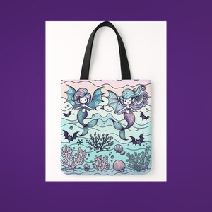 A pastel-colored tote bag features two cute mermaids with bat wings swimming in an underwater scene. Surrounded by coral, seashells, and playful bats, the background fades from soft pink to aqua. The mermaids have long flowing hair and heart details on their chests, giving a whimsical mix of spooky and sweet. The bag has black handles and is set against a purple and white backdrop