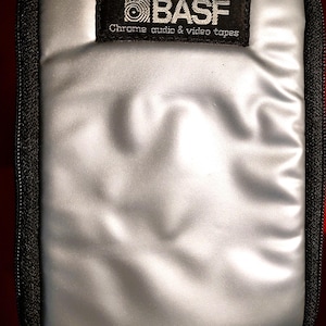 May include: A rectangular silver storage case with a black zipper border. A black fabric label is sewn onto the top center featuring the white text BASF and Chrome audio & video tapes. The silver surface has a smooth, slightly reflective finish.