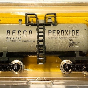 May include: A model train car with a gray tank labeled "BECCO PEROXIDE" and "BECK 895". The car has black wheels and a ladder. The model is in a clear plastic case with a yellow background. The word "ATLAS" is visible on the case.