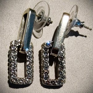 Pu&ograve; includere: Orecchini pendenti rettangolari color argento. Ogni orecchino presenta una cornice rettangolare incastonata con piccoli cristalli scintillanti. Gli orecchini hanno una chiusura a perno con un retro rotondo trasparente.