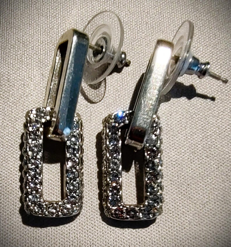Pu&ograve; includere: Orecchini pendenti rettangolari color argento. Ogni orecchino presenta una cornice rettangolare incastonata con piccoli cristalli scintillanti. Gli orecchini hanno una chiusura a perno con un retro rotondo trasparente.