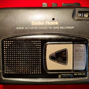May include: A black Radio Shack voice actuated cassette tape recorder featuring a speaker grille on the left, a central cassette compartment with a triangular window, a top-mounted tape counter, and a button labeled MIC POP-UP on the lower right.
