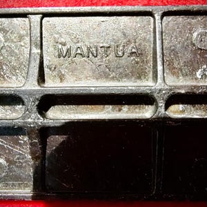 May include: A dark metallic rectangular component with a grid-like structural frame. The word MANTUA is embossed in capital letters in the center section. The surface shows a weathered, textured finish with visible casting marks and indentations.