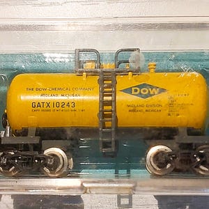 May include: A yellow model train tank car with the "DOW" logo and text, inside a clear plastic case. The tank car has black wheels and a ladder. A yellow label with text is in the case.