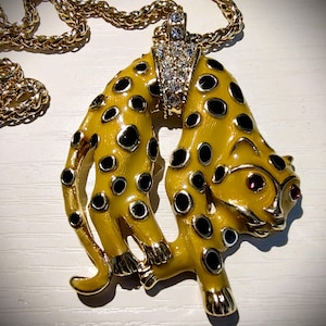 May include: A gold-toned necklace featuring a pendant in the shape of a stylized leopard. The pendant is yellow with black spots, gold accents, and red eye details. The necklace chain is also gold.