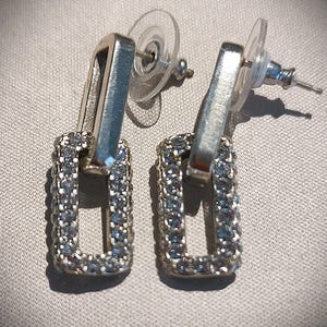 Pu&ograve; includere: Un paio di orecchini pendenti rettangolari argentati. Ogni orecchino presenta una cornice rettangolare incastonata con piccole pietre scintillanti. Gli orecchini hanno una chiusura a perno e farfalla.