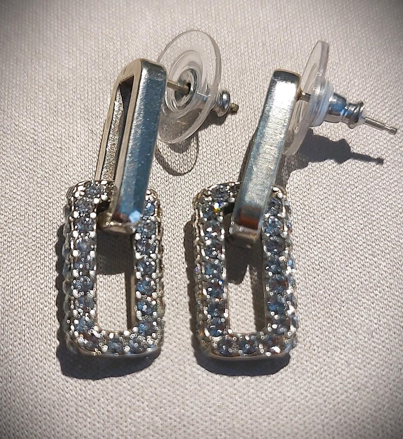 Pu&ograve; includere: Un paio di orecchini pendenti rettangolari argentati. Ogni orecchino presenta una cornice rettangolare incastonata con piccole pietre scintillanti. Gli orecchini hanno una chiusura a perno e farfalla.