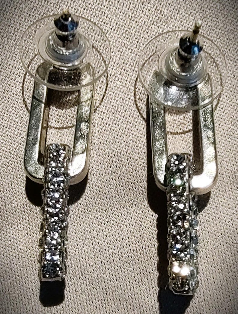 Pu&ograve; includere: Un paio di orecchini color argento con un design rettangolare pendente. La parte inferiore di ogni orecchino &egrave; incastonata con piccoli cristalli scintillanti. Gli orecchini hanno una chiusura a perno con un retro rotondo trasparente.