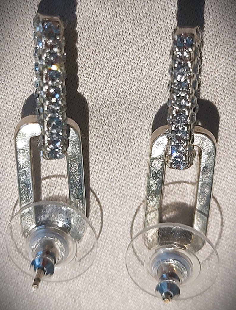 Pu&ograve; includere: Un paio di orecchini color argento. Ogni orecchino presenta una sezione superiore rettangolare incastonata con piccoli cristalli azzurri. La parte inferiore &egrave; un design a forma di U color argento. Sono attaccate chiusure per orecchini in plastica trasparente.