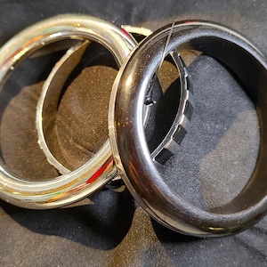 May include: Two bracelets are displayed. One is a wide, shiny silver bangle. The other is a dark, glossy, thick black bracelet. A thinner, silver bracelet with a geometric design is partially visible between the two.