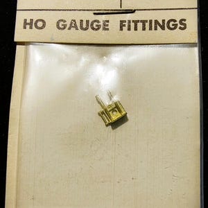 May include: A small gold-colored metal headlight bracket for model trains, packaged in a clear plastic bag attached to a beige cardboard header card. The card features the text CARY, HEADLIGHT BRACKET, HB-128, and HO GAUGE FITTINGS printed in black.