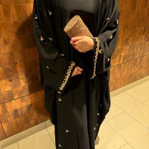 May include: A black abaya with gold embroidery and small gold embellishments. The abaya is paired with a gold clutch. The sleeves and edges of the abaya are decorated with gold detailing. The abaya is a traditional garment.