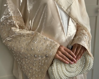 May include: A cream-colored abaya with a sheer overlay and beaded detailing on the sleeves. The abaya is paired with a white pearl clutch.