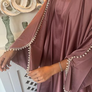 Puede incluir: Una kaftán de satén malva con ribetes de perlas alrededor de las mangas y el escote.