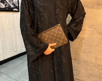 May include: A black abaya featuring tonal floral embroidery across the front and sleeves, paired with a brown and tan monogrammed clutch bag. The garment has long sleeves and a straight silhouette, while the clutch displays a repeating logo pattern.