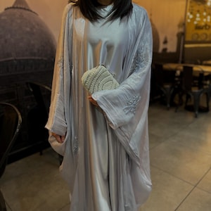 May include: A light gray, flowing abaya-style dress with intricate embroidery on the sleeves. The dress is paired with a matching shawl and a white beaded clutch. The fabric has a subtle sheen, suggesting a luxurious feel.