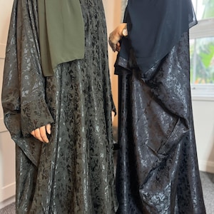 Puede incluir: Dos abayas negras largas con un sutil estampado floral. Una abaya es verde oscuro y la otra es negra. Ambas abayas tienen un corte holgado y fluido.