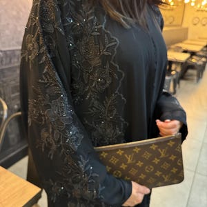 以下が含まれることがあります： 袖とフロントパネルに複雑な黒のフローラル刺繍と繊細なスパンコールの装飾が施された黒のアバヤ。金色のシンボルと文字が繰り返されるモノグラム柄の茶色のクラッチバッグを合わせています。