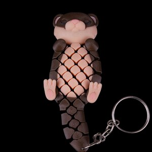 May include: A brown and pink 3D printed keychain shaped like a sleeping otter. The otter has a segmented body and a small metal ring attached to its tail.