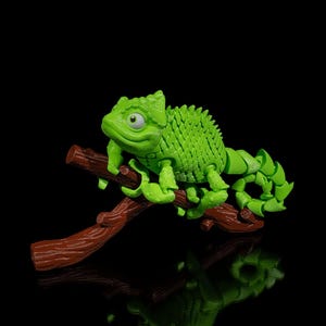 May include: A green chameleon toy with a textured, scaly body is perched on a brown branch. The chameleon has large, round eyes and a long, prehensile tail.