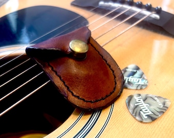 May include: A dark brown leather guitar pick holder with a brass snap closure, resting on the light wood body of an acoustic guitar. Two grey and white marbled Taylor brand guitar picks are positioned next to the leather pouch on the guitar.