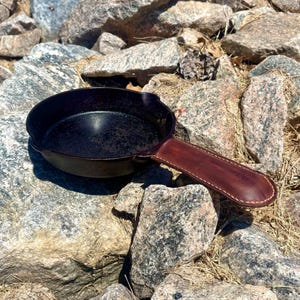 May include: A small, black cast iron skillet with a dark brown leather handle. The skillet is resting on a bed of gray and brown rocks. The handle is stitched with a light-colored thread.