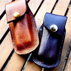 May include: Two rectangular leather pouches with rounded bottom edges and fold-over flaps. One pouch is a medium tan brown with a brass-toned metal snap button, and the other is a dark blackish-brown with a silver-toned metal snap button closure.
