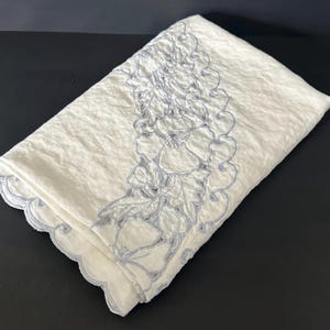 May include: Folded white linen cloth with a floral embroidered border in a light blue color. The embroidery features a scalloped edge and detailed floral patterns. The cloth is folded neatly, showcasing the design.
