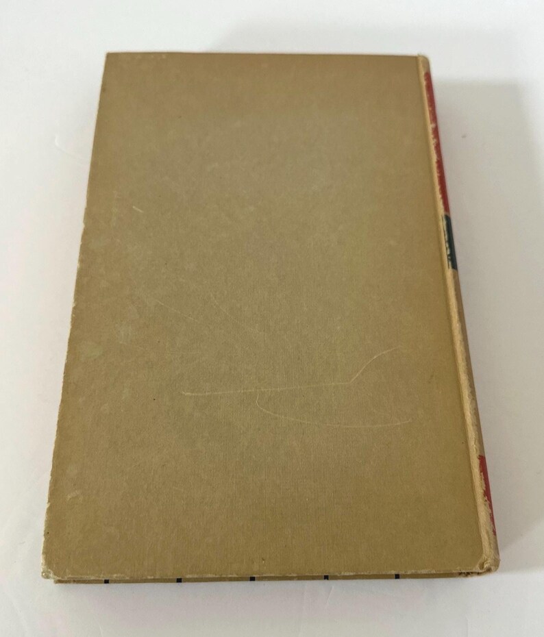 May include: A vintage book with a tan cover and a red, black, and blue spine. The cover shows signs of wear, with scratches and discoloration. The edges of the book are slightly worn, suggesting its age and use. The book's spine is visible, indicating the title and author.