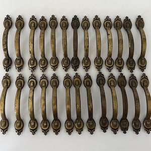 May include: A collection of twenty-six matching brass-toned metal drawer pulls arranged in two rows. Each handle features a curved grip with decorative, ornate mounting plates at both ends, displaying a weathered, aged finish throughout.