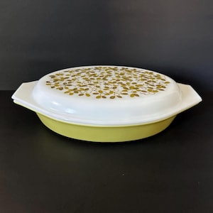 May include: A vintage Pyrex oval casserole dish with a white lid featuring a gold and olive green floral pattern. The dish itself is a pale yellow-green color. The dish has handles on either side.