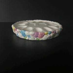 May include: White ceramic Easter egg holder with a scalloped edge. The holder has multiple egg-shaped indentations and is decorated with colorful bunnies and flowers around the rim. The bunnies are in shades of blue, purple, and yellow.