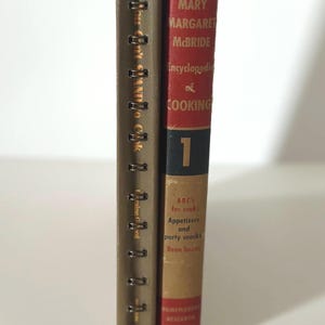 May include: Two vintage cookbooks stand side-by-side. One is bound with metal rings and has text in a foreign language. The other, titled "Encyclopedia of Cooking," has a red spine with the number 1 and author's name.