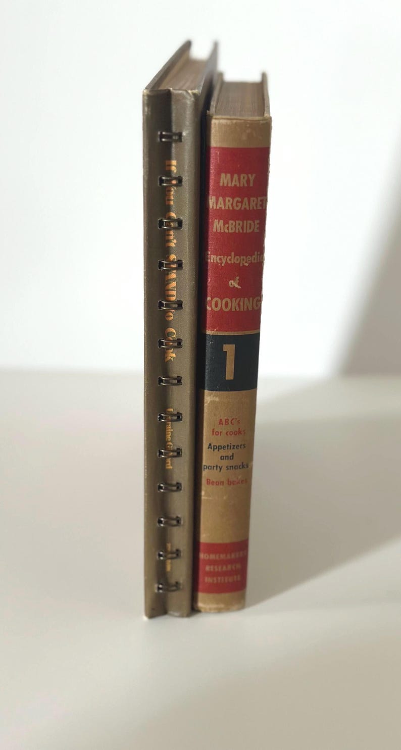 May include: Two vintage cookbooks stand side-by-side. One is bound with metal rings and has text in a foreign language. The other, titled "Encyclopedia of Cooking," has a red spine with the number 1 and author's name.