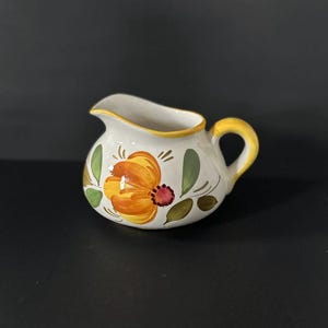 May include: A white ceramic creamer with a yellow, orange, and green floral design. The creamer has a handle and a rounded body.