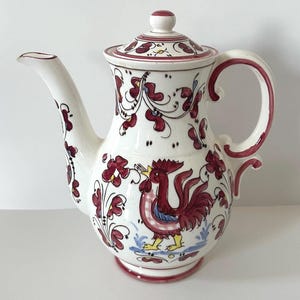 May include: White ceramic coffee pot with a rooster design. The pot features a red and white floral pattern, a curved handle, and a lid with a matching design. The rooster is depicted in red, blue, and yellow.