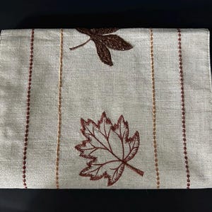 May include: Rectangular beige placemat with embroidered brown fall leaves and vertical stripes. The placemat features two large leaves and two vertical lines of brown stitching, adding a touch of autumn to any dining table.