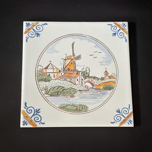 May include: Square ceramic tile featuring a Dutch windmill scene in a circular frame. The windmill has orange and brown tones, with a bridge and water in the foreground. Decorative blue and orange corner accents complete the design.