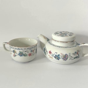 May include: A white ceramic teapot and matching teacup featuring a delicate floral pattern in shades of blue, pink, and green. Both pieces have a rounded shape, a single handle, and a thin metallic gold rim accenting the edges.